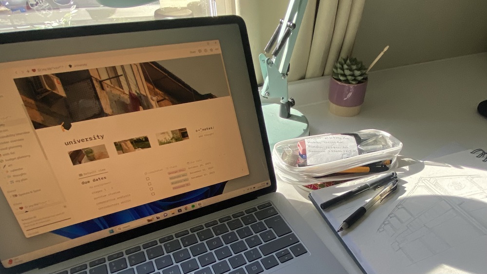 A laptop computer open on a desk, showing university work on screen. Also on the desk is an architectural drawing.