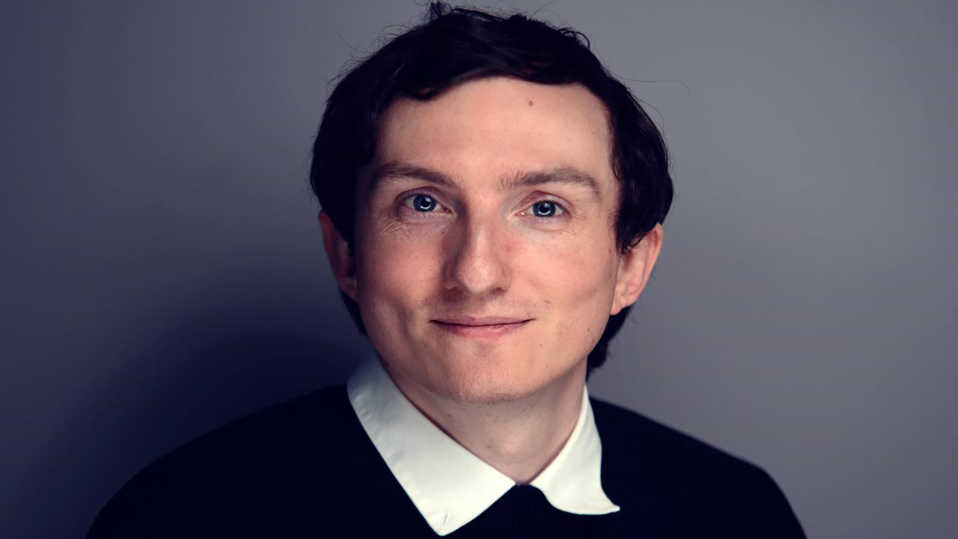 A headshot of David McCallig smiling, while wearing a white shirt and black jumper