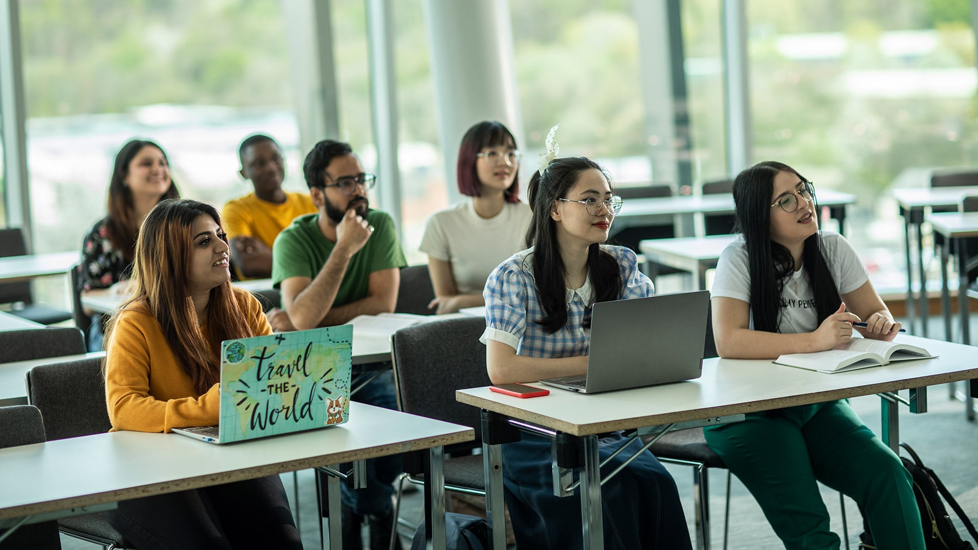 A group of seven international students sat at desks, looking towards their teacher.
